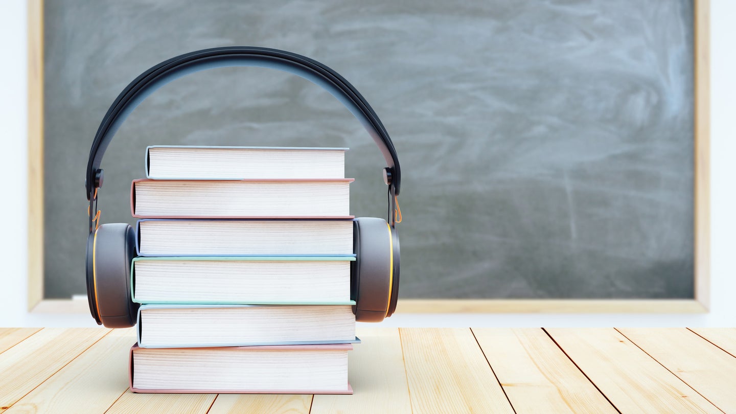 A stack of six hardcover books on a wood table with headphones around them and a blackboard in the background.